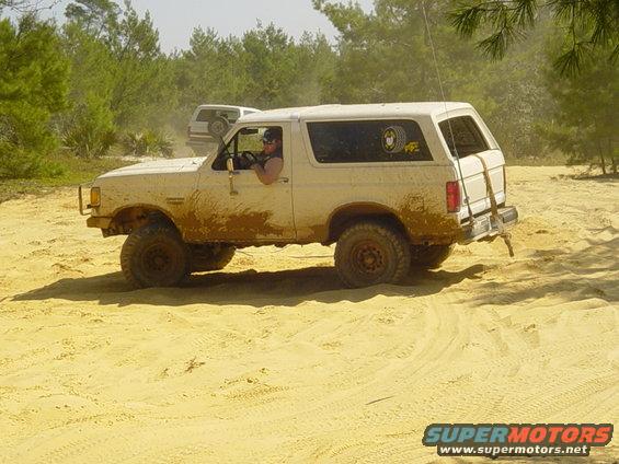 White Ford Bronco driving on a sandy trail, covered in mud. Another Bronco is behind it