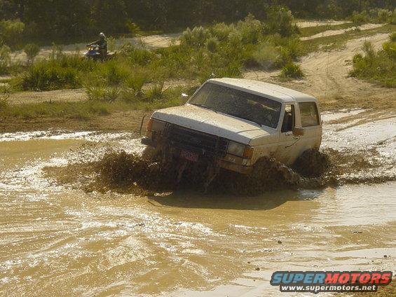 1989 Ford Bronco driving through a mud puddle, kicking up water and mud