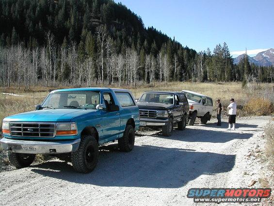 Three Ford Broncos parked on a dirt road with a wooded hillside in the background. The lead Bronco is teal
