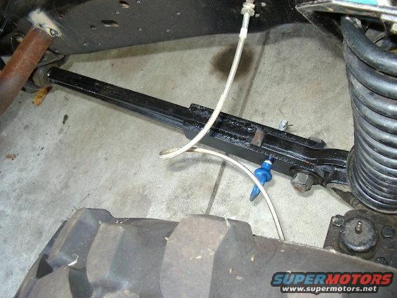 Black metal suspension arm with brake line and coil spring attached, near a large tire