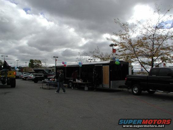 Black pickup truck parked next to a black trailer with balloons. Other vehicles and people are visible in the background