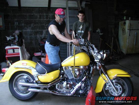 Man in red hat and denim shorts with a yellow cruiser motorcycle. Another man stands behind him