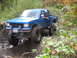 1990 Ford Bronco - A blue pickup truck tackles a challenging muddy trail with its large off-road tires.