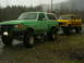 1990 Ford Bronco - Two lifted 4x4s, a green Bronco and a yellow F-Series, sit parked on a gravel lot during a rainy day.