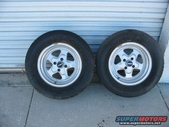 Two tires mounted on silver five-spoke wheels, resting on concrete in front of a corrugated metal door