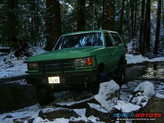 1990 Ford Bronco driving through a snowy forest with trees and rocks