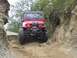 1990 Ford Bronco - A red Jeep Wrangler navigates a narrow, rocky off-road trail with steep dirt banks on either side.