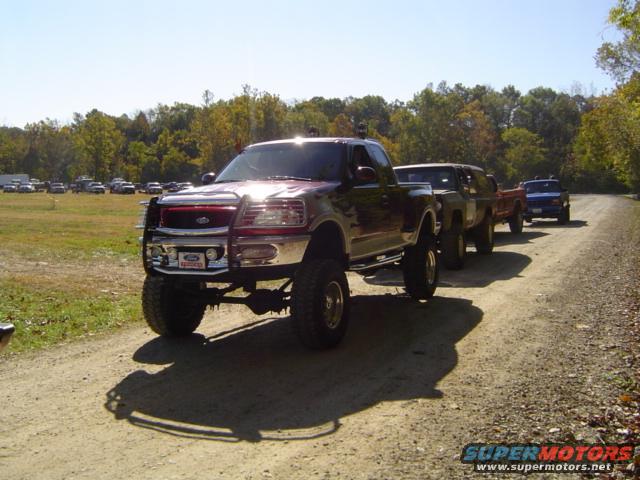 Several lifted pickup trucks are lined up on a dirt road. The lead truck is a dark red Ford F-150