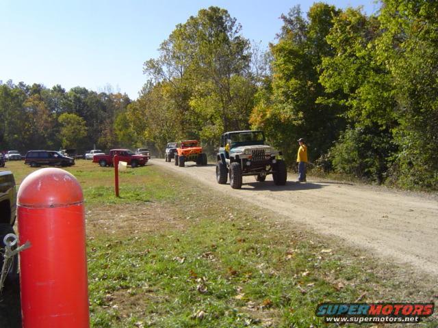 Green Jeep Wrangler with large tires on a dirt road, with other vehicles parked in the background
