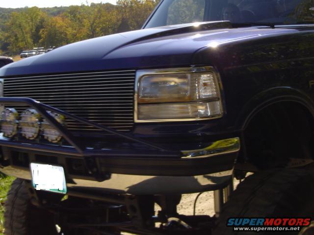 Front end of a dark blue Ford F-Series truck with a chrome grille and aftermarket bumper with lights. License plate is obscured