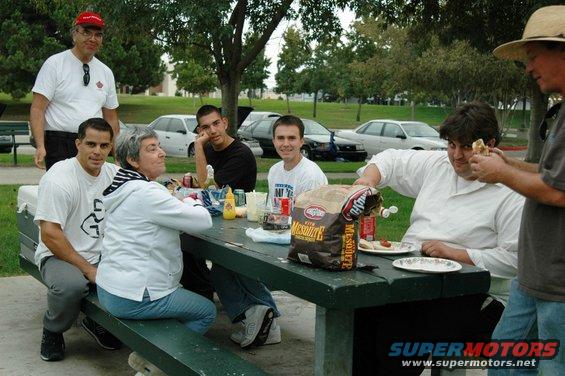 People gathered around a picnic table with cars parked in the background