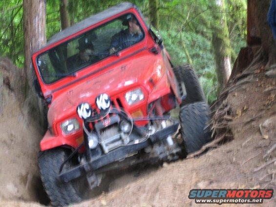Red Jeep Wrangler climbing a steep dirt hill with trees around. Has KC lights and a winch