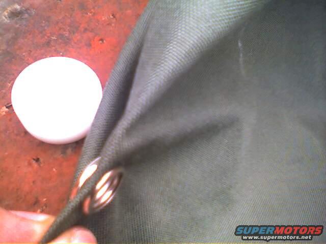 Close-up of green fabric with metal grommets, next to a white round object. Supermotors.net logo visible