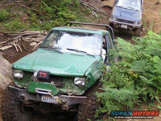 Green off-road truck with winch and damaged front end, blue Toyota in background on trail