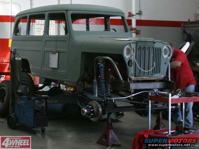 Green Jeep Willys Wagon front view, undergoing restoration with exposed chassis and suspension