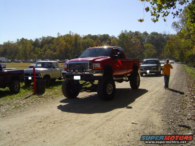 Lifted red pickup truck with large tires on a dirt road, other trucks and trees in background