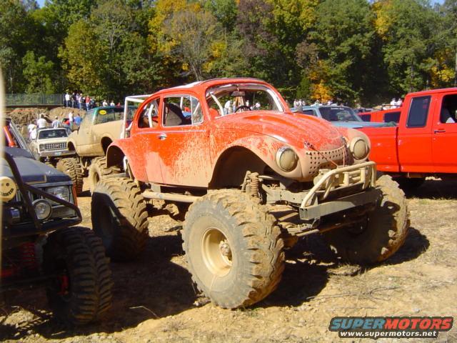 Orange Volkswagen Beetle modified for mud bogging, with large tires and roll cage. Other trucks and vehicles are visible in the background