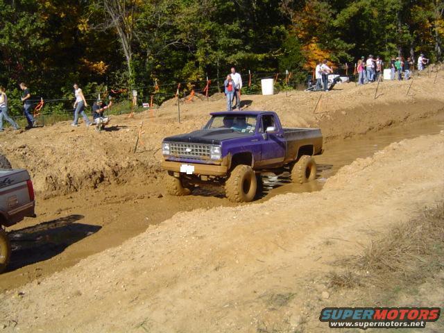 Purple pickup truck with lifted suspension and large tires driving through mud, front grille visible