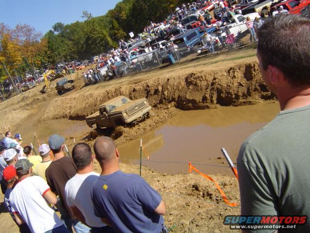 Muddy truck stuck in a bog pit with spectators watching. Another truck is in the background