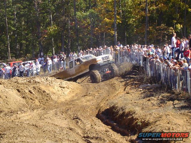 White off-road truck with large tires climbing a dirt hill, crowd watching behind a fence