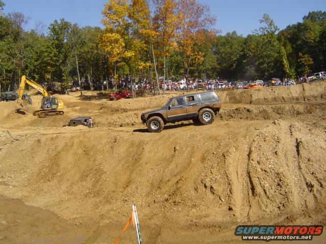 Brown SUV with large tires on a dirt hill, with spectators and excavators in the background