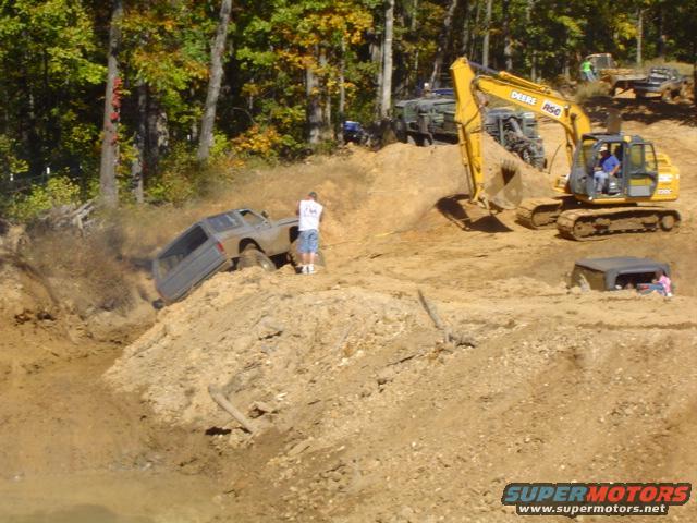 Off-road vehicles struggling in a mud pit, with a yellow excavator nearby. One vehicle is partially submerged