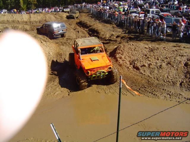 Orange off-road vehicle navigating a mud pit with spectators watching from a berm