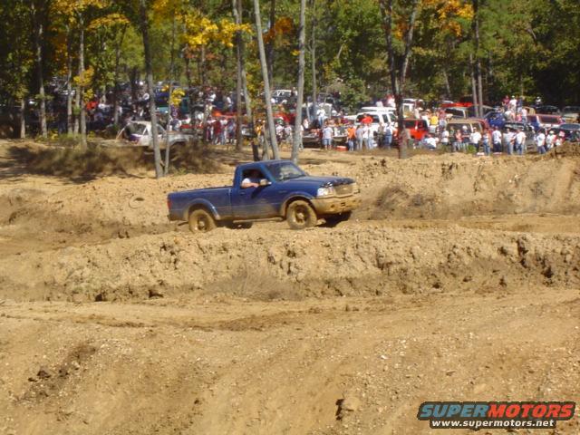 Blue pickup truck driving through a muddy track with trees and spectators in the background