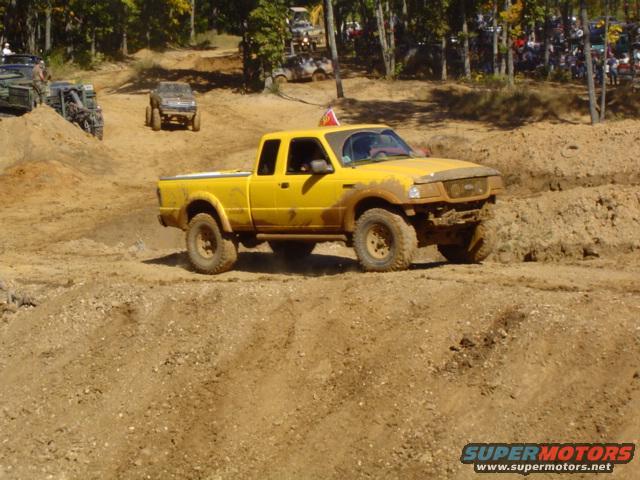 Yellow pickup truck driving on a dirt track with other vehicles and spectators in the background
