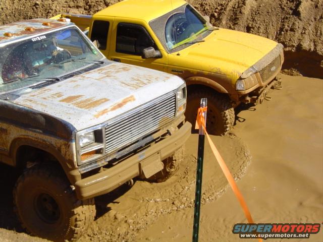 Mud-splattered white truck front grille and headlight, next to a yellow truck in mud