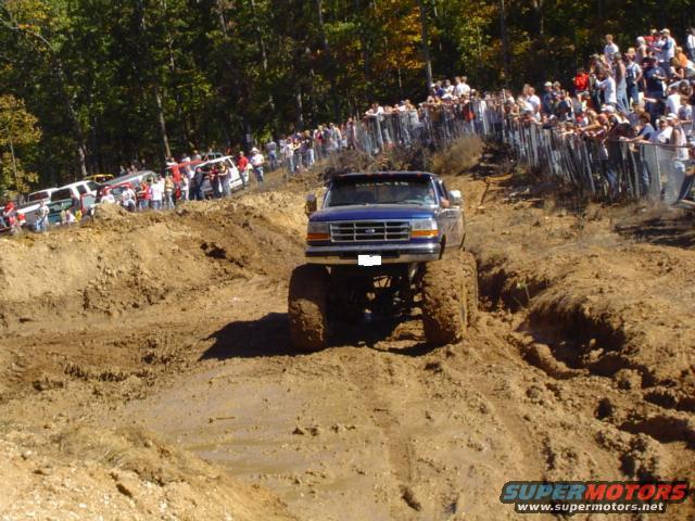 Blue lifted pickup truck with large tires driving through mud, crowd watching