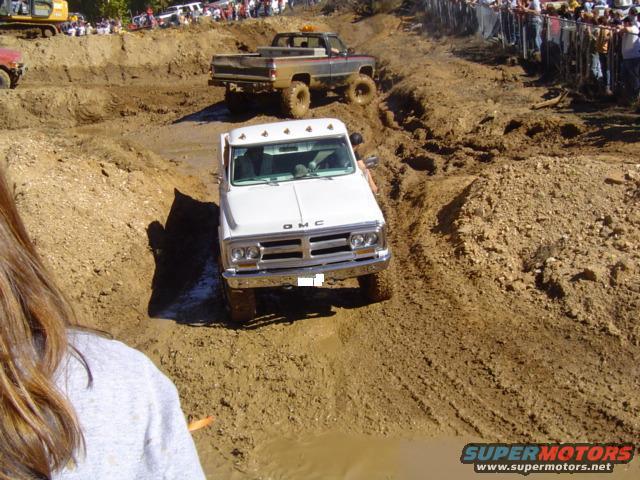 White GMC pickup truck driving through a mud pit with a brown pickup truck behind it and spectators watching