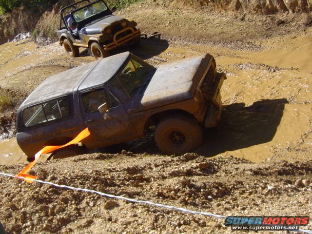 Muddy dark SUV stuck in mud, orange strap attached to front, another Jeep behind it