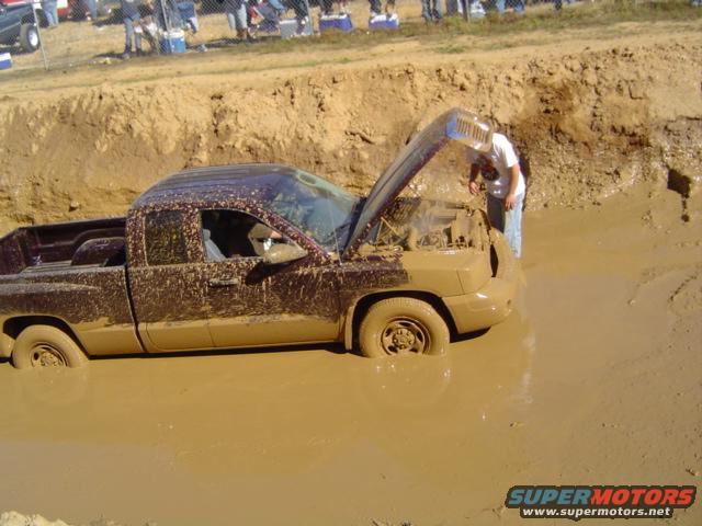 Mud-covered pickup truck stuck in a mud pit with hood open, person looking at engine