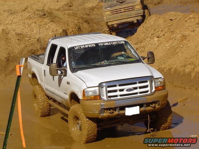 White pickup truck with "LIFT EM UP BOYS FAT GIRLS CANT CLUB" sticker on windshield, driving through mud