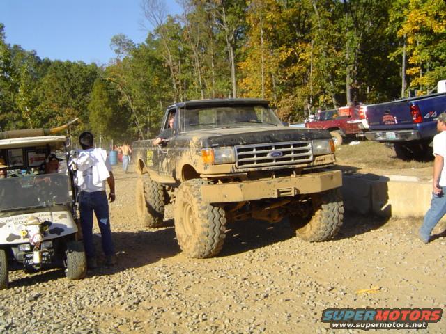 Mud-covered black pickup truck with large tires, front view. Grille suggests 1980s model