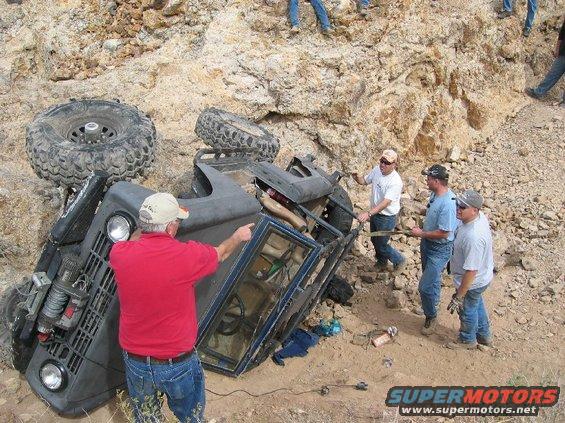 Overturned dark blue off-road vehicle on rocky terrain, with people attempting to right it