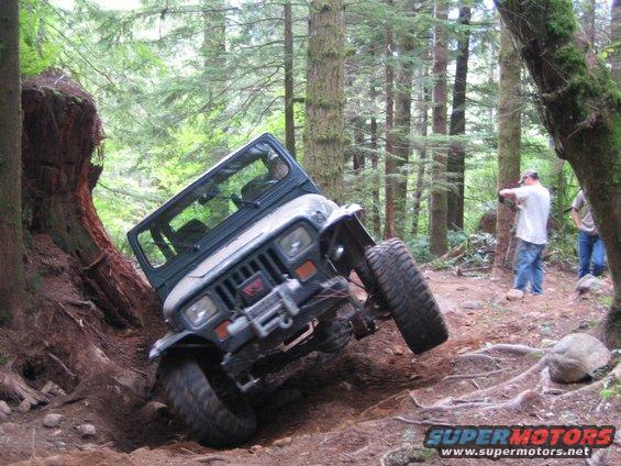 Jeep Wrangler tilted on its side on a dirt trail in a forest, with two people watching