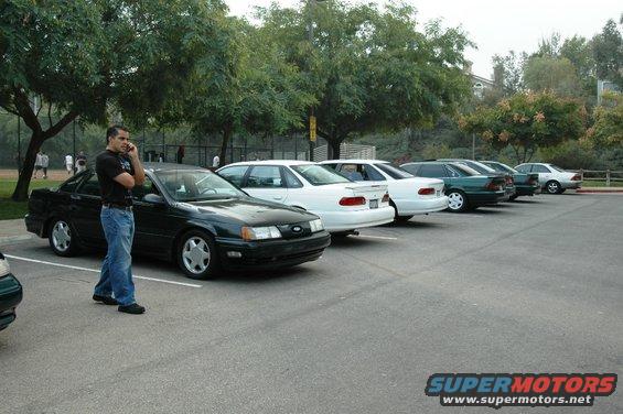 Man on phone next to black 1991 Ford Taurus, parked in a line with other sedans
