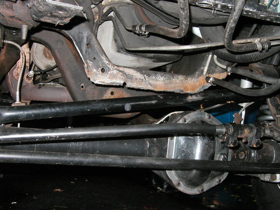 Underside view of vehicle suspension and drivetrain components, showing rusty frame and metal parts