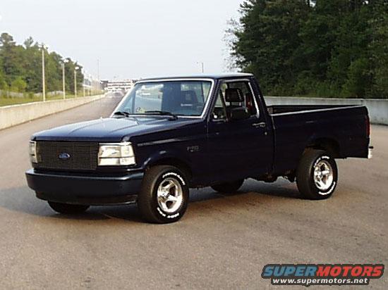 Dark blue Ford F-150 pickup truck parked on asphalt, trees and a track in background