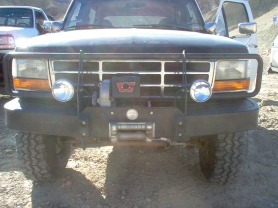 Black Ford Bronco with winch and brush guard, front view. White truck visible to the left
