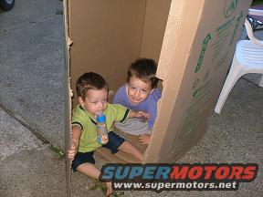Two young kids playing inside a large cardboard box. One has a bottle