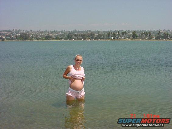 Pregnant person in shallow water, city skyline and trees in background. Watermark reads SUPERMOTORS