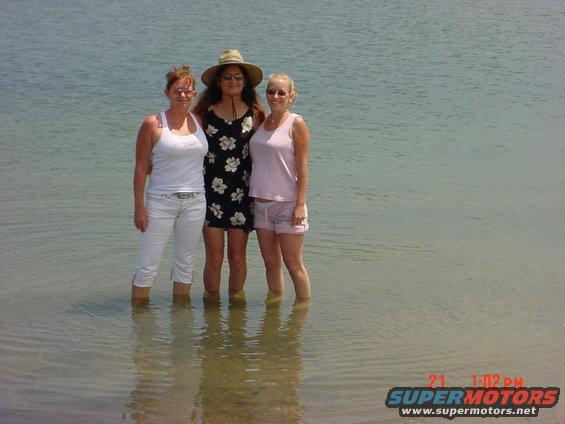 three women standing in shallow water, one wearing a hat and floral dress, another in a white tank top and pants, and the third in a pink tank top and shorts