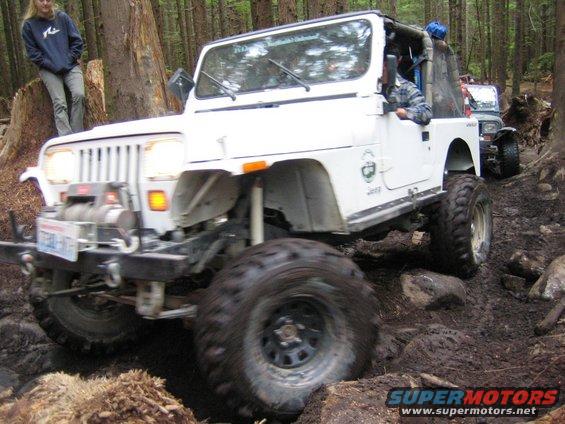 White Jeep Wrangler driving on a muddy, wooded trail. Another Jeep is visible behind it