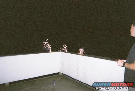 Person holding a sparkler near fireworks on a white railing at night