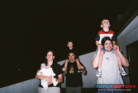 Family members, including adults carrying children on their shoulders, look up at the night sky