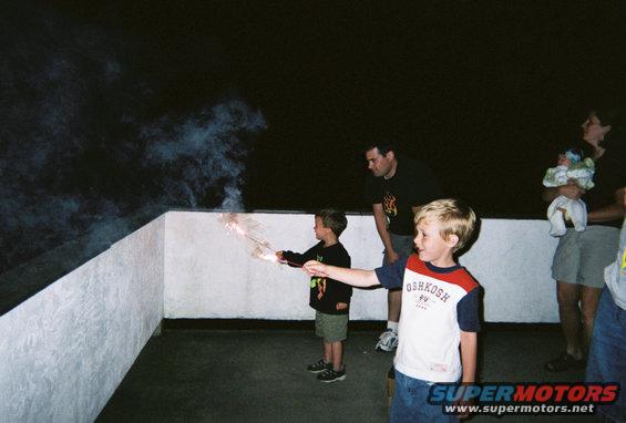 Children holding sparklers on a balcony at night, with adults in the background