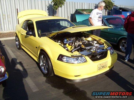Yellow Ford Mustang with hood and trunk open, showing engine and interior. California license plate reads SUZNNOL